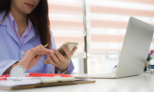 Female clinician using smartphone, sitting in front of laptop